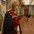 Schaufensterpuppe im Franziskanermuseum trägt mittelalterliche Frauenkleidung: rotes Gewand, blauer Mantel mit silberner Spange und Kopftuch; steht im Museumsflur neben Vitrinen und Schilden.