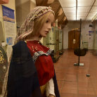Lebensgroße Frauenfigur in mittelalterlicher Kleidung: rotes Kleid, bestickter Kragen, helles Kopftuch mit Schleier, Ausstellungsobjekt im Flur des Franziskanermuseums, Blick nach unten.