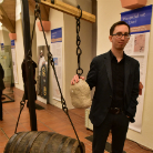 Im Museum steht ein Mann neben einer historischen Balkenwaage mit hängendem Sack als Gewicht; im Vordergrund ein liegendes Holzfass, im Hintergrund Vitrinen und Informationstafeln.