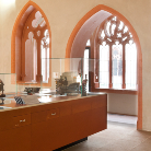 Heller Museumsraum des Franziskanermuseums mit zwei gotischen Spitzbogenfenstern und Maßwerk; im Vordergrund eine orangefarbene Vitrine mit Ausstellungsobjekten