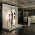 Ausstellungsraum des Franziskanermuseums: beleuchtete Glasvitrine mit lebensgroßem Schaufensterpuppe in franziskanischer Kutte, dahinter gerahmte Bilder; weitere Vitrinen mit Exponaten im dunklen Raum.