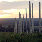 Skulptur aus schlanken, unterschiedlich hohen vertikalen Holzpfählen auf einem Grashügel, dahinter Wald und untergehende Sonne.