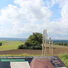Blick von einer Aussichtsplattform mit Informationstafel und mehreren schlanken Holzstelen über weite Felder, einen einzelnen Baum und weiten, bewölkten Himmel