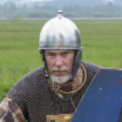 Porträt eines Mannes in mittelalterlicher Rüstung mit Kettenhemd, silbernem Helm und Schild vor offener Wiese