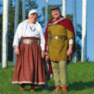 Zwei Menschen in historischen Gewändern — eine Frau mit weißem Kopftuch und rotem Rock, ein Mann mit grüner Tunika, rotem Umhang und Mütze — stehen nebeneinander auf einer grünen Wiese vor schlanken weißen Birken.