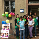 Neun lächelnde Personen unterschiedlichen Alters stehen vor dem Eingangsportal des Franziskanermuseums; rechts eine Tür, links bunte Luftballons und ein Informationsaufsteller mit Bildern.