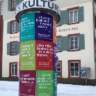Zylindrische, bunte Litfaßsäule mit verschiedenfarbigen Textfeldern zu Kulturveranstaltungen, steht auf schneebedecktem Gehweg vor einem historischen Gebäude