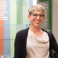 Lächelnde Person mit kurzen blonden Haaren und Brille, trägt helles Oberteil und dunkle Strickjacke, steht vor farbigen Ausstellungstafeln im Museum.