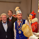 Gruppe bei Fasnachtsfeier: Mann in blauem Festgewand mit goldener Ordenkette und hoher Federhaube hält einen verzierten Stab; neben ihm zwei Personen in roten Narren-Kostümen und zwei Männer in dunklen Anzügen, alle lächelnd