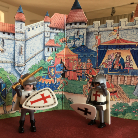 Zwei mittelalterliche Ritterfiguren vor bemaltem Burgpanorama: links Ritter mit weißem Schild und rotem Kreuz sowie Lanze, rechts Ritter mit schwarzem Kreuz, beide mit Helmen und Rüstungen; im Hintergrund farbige Burg- und Zeltdarstellung.