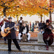 Historisch kostümierte Straßenmusikanten vor dem Franziskanermuseum: Gitarrist, Violinistin und Schlagzeuger spielen auf Treppen, Publikum im Hintergrund, goldene Herbstblätter an den Bäumen.