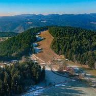 Luftaufnahme einer Hügellandschaft: bewaldete Höhenrücken, lichtes Wiesental mit Schneeresten und entfernten blauen Bergen unter klarem Himmel.