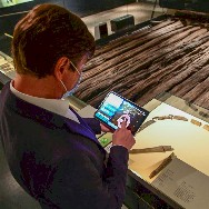 Ein Besucher mit Mund-Nasen-Schutz im Franziskanermuseum betrachtet ein Tablet; neben ihm liegt ein aufgeschlagenes historisches Buch und ein langes hölzernes Ausstellungsstück.