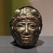 Bronzener antiker Kopf als Maskenrelief: männliches Gesicht mit Helm, schmale Augen, gerade Nase, geschlossener Mund und grünliche Patina