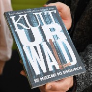 Nahaufnahme: Hände halten ein Heft mit dunkelblauem Umschlag und großem weißen Titel KULTURWALD, darunter ein kleiner Untertitel, ein orangefarbener Akzent am Cover.