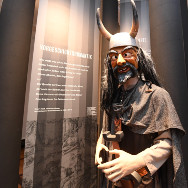 Lebensgroße Figur eines bärtigen Kriegers mit Hornhelm und langem dunklem Haar, braunem Umhang, die eine Axt hält; im Hintergrund Museumstafeln.