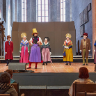 Theateraufführung in Kirchenraum: sieben Darsteller in bunten historischen Kostümen und Hüten auf einer Holzbühne, dahinter hohe Bogenfenster; Publikum sitzt in Kirchenbänken.
