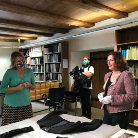 Drei Personen im Magazin des Franziskanermuseums untersuchen auf einem Tisch ausgelegte historische Textilien; Regale mit Büchern und Ordnern, Beleuchtung und Kameraausrüstung im Raum.