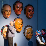 Ausstellungsraum mit sechs großen geschnitzten Narrenköpfen an der Wand; davor zwei lebensgroße kostümierte Figuren, eine hält eine kleine Maske.