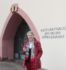 Eine Frau in roter Jacke und buntem Schal steht vor dem rosafarbenen Spitzbogenportal des Alten Rathauses mit der Beschriftung Altes Rathaus Museum Bürgeramt und hält eine Kamera.
