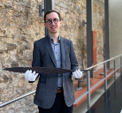 Museumspersonal mit weißen Handschuhen hält vorsichtig ein langes, schmales Holzbrett; steht vor alter Sandsteinmauer und Metallgeländer im Ausstellungsraum.