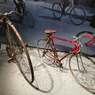 Zwei historische Fahrräder in einer Museumsausstellung: rechts ein rotes Rennrad mit gebogenem Lenker und braunem Ledersattel, links ein älteres, rostiges Velo mit großem Vorderrad; Schatten weiterer Fahrräder an der Wand.