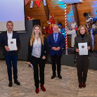Gruppenfoto bei einer Auszeichnung: mehrere Personen stehen in einem Saal, drei von ihnen halten Urkunden; im Hintergrund Holzvertäfelung und bunte Girlanden.