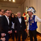 Im Museum: Ein Museumsführer in bunter historischer Tracht mit großem Federhut erläutert drei Gästen in formeller Abendkleidung vor Exponaten.
