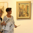 Zwei Besucherinnen betrachten im Museum eine gerahmte Altstadt-Straßenmalerei; sie halten Informationsblätter, im Hintergrund weiße Wand und eine weitere gerahmte Arbeit.