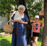 Frau in historischer franziskanischer Kleidung (blaues Gewand, weißes Kopftuch) mit Korb steht neben einem Mädchen in Sommerkleidung und Mütze vor einem Baum im Freien