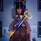 Zwei lebensgroße Figuren in blauen Gewändern hängen an einem übergroßen goldenen Schlüssel vor der Fassade eines historischen Hauses mit verziertem Portal.