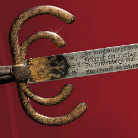 Nahaufnahme einer historischen Degenklinge: dreigeschwungener, vergoldeter Handschutz mit Patina und eine Stahlklinge mit feiner eingravierter Schrift.
