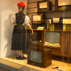 Schaufenster des Franziskanermuseums: Mannequin in schwarzem Dirndl mit rotem Hut neben einem Sideboard im Mid-Century-Stil und zwei alten Röhrenfernsehgeräten, Regalfächern mit Radiogeräten und Dekor — Ausstellung einer 1950er/60er-Jahre-Wohnzimmerszene.