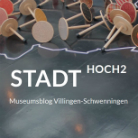 Mehrere hölzerne Kreisel mit Stielen liegen auf dunklem Untergrund über einer weiß aufgemalten Stadtsilhouette; darüber der Schriftzug STADT HOCH2 und kleiner Hinweis Museumsblog Villingen-Schwenningen