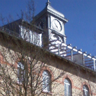 Teilansicht eines historischen Backsteingebäudes des Franziskanermuseums mit drei Rundbogenfenstern; darüber weißer Uhrturm mit rundem Zifferblatt und Brüstung, blauer Himmel und kahler Baum im Vordergrund