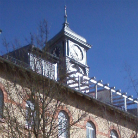 Backsteingebäude mit drei Bogenfenstern, darüber ein weißer Uhr- und Glockenturm mit sichtbarem Zifferblatt; blauer Himmel und kahle Baumzweige im Vordergrund.