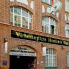 Rote Backsteinfassade eines historischen Fabrikgebäudes mit großen Rundbogenfenstern; schwarzes Schild Württembergische Uhrenfabrik über einem Tor, darunter der Eingang zum Uhren-Industrie-Museum.