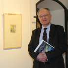 Älterer Mann in dunklem Anzug und Krawatte steht im Franziskanermuseum neben einem gerahmten kleinen Bild an der Wand und hält mehrere Broschüren.