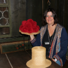 Eine sitzende Frau hält eine große rote Filzblume als Hutverzierung; vor ihr steht ein Strohhut auf einem Holztisch. Dunkler Museumsraum mit Holzbank und bleiverglastem Fenster im Hintergrund.