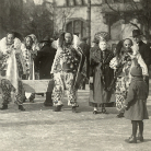 Historische Schwarzweißfotografie einer Fastnachtsgruppe in traditionellen Kostümen und Holzmasken; mehrere verkleidete Personen stehen auf einem Platz vor einer Gebäudefassade.