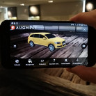 Eine Hand hält ein quer gehaltenes Smartphone, dessen Bildschirm eine gelbe Auto-3D‑Modellgrafik in Augmented Reality auf einem Holzboden zeigt; App-Bedienelemente am Bildschirmrand sichtbar.