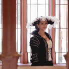 Frau in schwarzer historischer Tracht mit weißer Federhaube und Zöpfen, stehend in einem Saal mit hohen Fensterbögen und Säulen, Blick zur Seite.