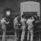 Schwarzweißfoto: Vier Männer in Arbeitskleidung bedienen eine historische Schalt- bzw. Vermittlungsanlage mit zahlreichen Steckverbindungen; links an der Wand hängt eine Uhr.