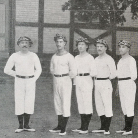 Schwarzweißfoto: Sechs junge Männer in weißen Sportuniformen mit Gürtel, Kopfband und dunklen Kniestrümpfen stehen nebeneinander vor einer Hauswand.