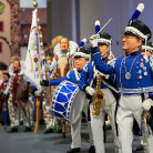 Festumzug mit Personen in historischen blau-weißen Uniformen; vorne ein junger Trommler, dahinter Fahnenträger und weitere Teilnehmer in traditionellen Gewändern.