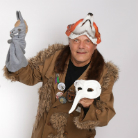 Person in brauner, mit Pelz besetzter Jacke hält in einer Hand eine weiße Halbmaske, in der anderen eine graue Handpuppe mit Zipfelmütze; trägt bunte Mütze und lächelt vor hellem Hintergrund.
