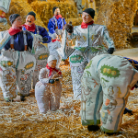 Gruppe maskierter Personen in bemalten, voluminösen Stoffkostümen mit Gesichter- und Tiermotiven; Erwachsene und ein Kind stehen auf Strohboden vor Heuballen in einer traditionellen Festszene