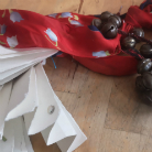 Rotes Stoffband mit einer Kette aus Metallglöckchen, daneben ein gefaltetes weißes Papierornament auf Holzboden.