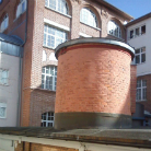 Zylindrische rote Backsteinstruktur mit flachem Dach und Metallsockel vor einem historischen Backsteingebäude mit mehreren Fenstern